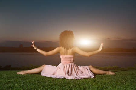 Back view of flexible dancer sitting on grass at sunset
