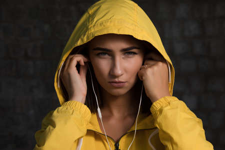 Serious woman under yellow hood with earbuds listen a musicの写真素材