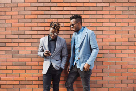 Two african men friends ready to call boss by smartphone, brick wall backgroundの写真素材