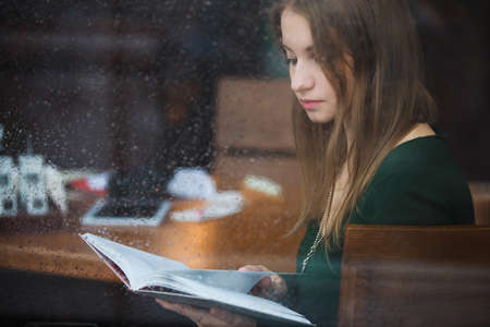 Woman reading book in the cafe, view through wet window at rainy dayの写真素材