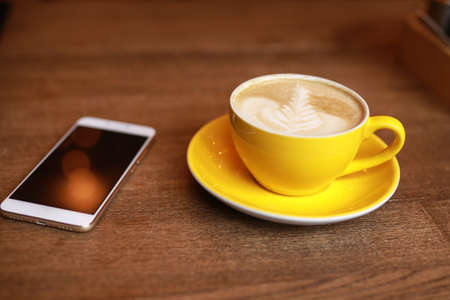 Coffee-break, latte in a yellow cup and smartphone on a deskの写真素材