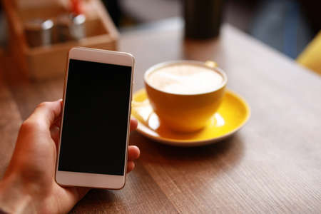 Coffee-break, male hand holding empty screen of smartphone in cafe, yellow cup with latte on...