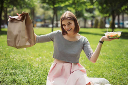 Woman holding and demonstrating fresh salad sitting at summer park, healthy food concept, empty blank paper bagの写真素材