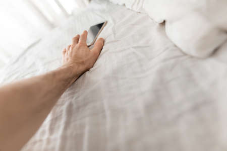 Male hand reaches to the smartphone immediately after wake up in bedの写真素材