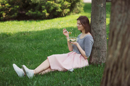 Woman eating fresh salad sitting at summer park leaning on tree trunk, healthy food conceptの写真素材