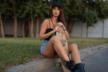 Young stylish skateboarder woman sitting with her skateboard on the roadの写真素材