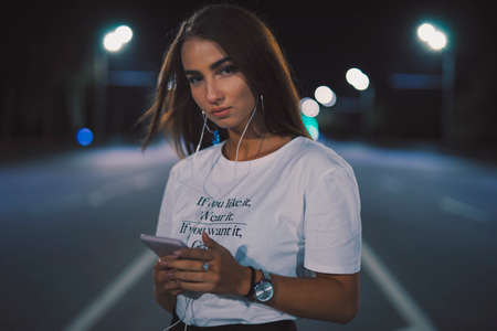 Portrait woman in earbuds at night road listening to music in earphones standing enjoying playlist songs on mobileの写真素材