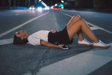 Portrait woman in earbuds lying on night road listening to music in earphones enjoying playlist songs on mobileの写真素材