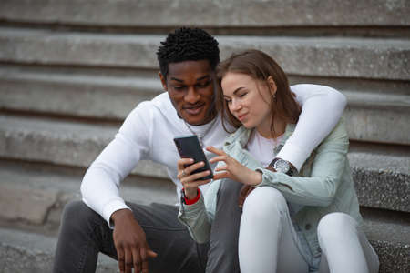 Two multiracial friends tourists looks photos in smartphone sitting in a cityの写真素材
