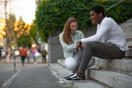 Two multiracial friends African man and Caucasian woman talking sitting in a cityの写真素材
