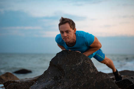Young strong man doing push-ups from stone at beach working out exercise with ocean background