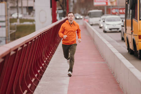 Young athlete man in orange hoodie running fast on the bridge