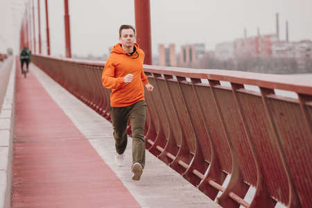 Young athlete man in orange hoodie running fast on the bridge