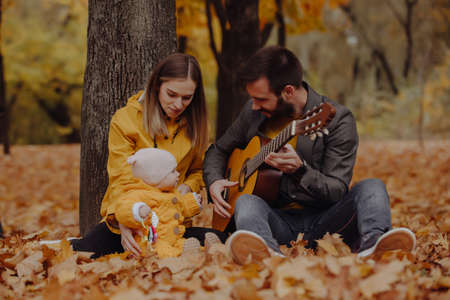 Happy young family sitting with little baby at the autumn park, dad plays on guitarの写真素材