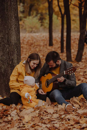 Happy young family sitting with little baby at the autumn park, dad plays on guitarの写真素材