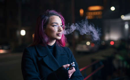 Unconventional woman with pink hair smoking electronic cigarette in night cityの写真素材