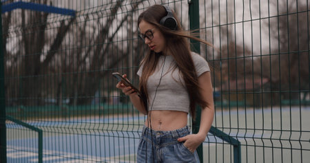 Girl standing near fence on a stadium in a park wearing headphones listening to music in smartphoneの写真素材