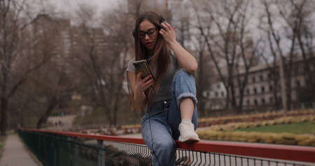 Happy girl listening to music in headphones sitting on fence in a parkの写真素材