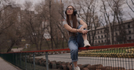 Happy girl listening to music in headphones sitting on fence in a parkの写真素材