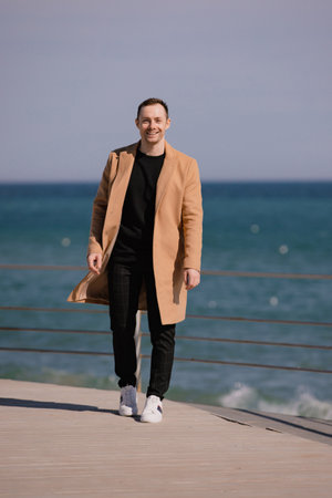Stylish man in the beige coat walking along sea shoreの写真素材