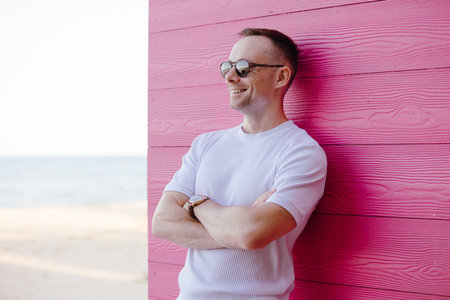 Handsome fashion smiling man posing with sunglasses leaning on pink wooden wall in the white costume, arms crossedの写真素材