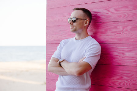 Handsome fashion man posing with sunglasses leaning on pink wooden wall in the white costume, arms crossedの写真素材