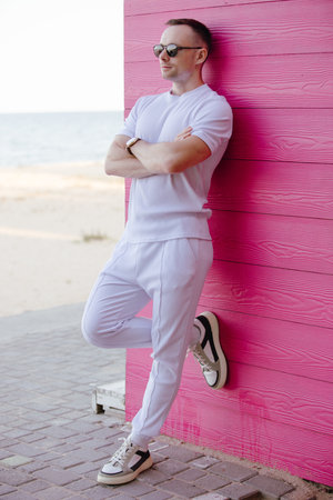 Handsome fashion man posing with sunglasses leaning on pink wooden wall in the white costumeの写真素材