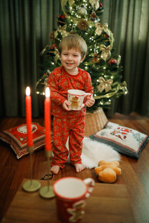 Little boy in red pajama sitting near the Christmas tree at home holding cupの写真素材