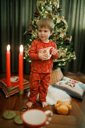 Little boy in red pajama sitting near the Christmas tree at home holding cupの写真素材