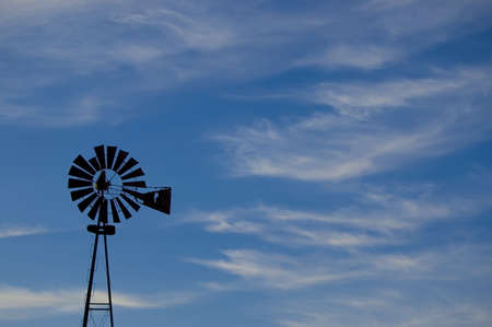 Silhouetted windmill with blue, cloudy sky. Old and rusty, with rural charm.の写真素材