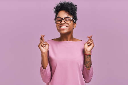 I have to win. Portrait of young female has dark skin, clenches teeth, raises fingers crossed, makes desirable wish, wears round spectacles and casual clothes, waits for good newsの写真素材