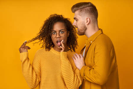 Handsome ginger man whispering secret or interesting gossip to young black woman in her ear isolated over yellow background. African girl feels amazed to hear confidential information from companion.の写真素材