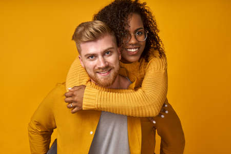 Young red-haired man piggybacking his dark skinned girl looking with happy expression at camera isolated over orange background. Two friends with specific appearance making joy.の写真素材