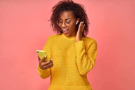 Overjoyed happy excited Afro American female glad to answers the video call using wireless earphones, rejoices good news, stares at the mobile phone, celebrates her success. Technology concept.の写真素材