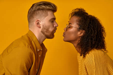Mixed race couple stand in profile, keep lips folded, staring at each other, going to kiss, dressed casual mustard color wear, make grimace, pose over orange wall, have fun. Facial expressions conceptの写真素材