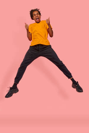 Happy satisfied black man is jumping in the air and shows with both hands approval gesture, keeps two thumbs raised, smiles broadly, wears yellow t-shirt, against pink wall. Thats nice, I like itの写真素材