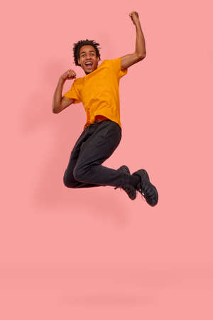 Full length photo of cheerful African American man jump raise fists to scream yes celebrate a victory or rejoicing with sales starting, wear orange casual t-shirt isolated over pink backgroundの写真素材