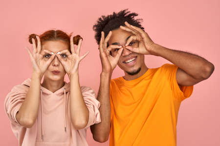 Funny african-american male and ginger caucasian female holds hands near eyes, pretends to look through binoculars tries to find love good offer, shows zero or okay gesture over eyes, on pink wallの写真素材
