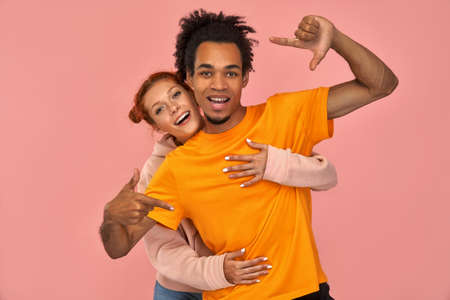 Smiling young dark skinned man with cheerful expression, makes hand frame, tries to find right perspective, searches inspiration or right angle, his ginger girlfriend hugging behind, pink backgroundの写真素材