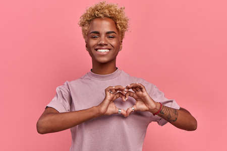 Portrait of happy african woman confesses in love, shows heart gesture, smiles broadly, has romantic...