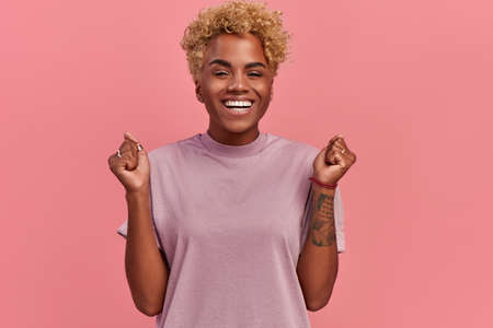 Portrait of overjoyed black female clenches fists with happiness, opens mouth widely as shouts loudly, celebrates her success, poses against pink background. Happy African woman with joyful expressionの写真素材
