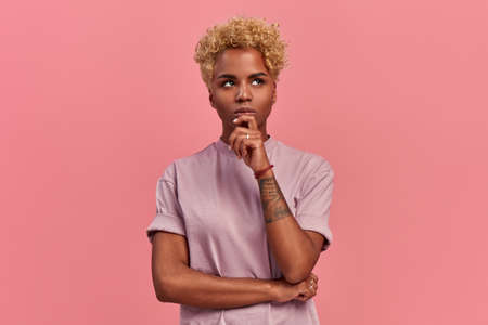 Pensive attractive curly blonde African American female being deep in thoughts, looking up, keeps forefinger near the chin, wears fashionable lilac t shirt, stands against a pink background.の写真素材