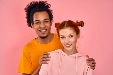Close-up positive beautiful mixed race couple charming red-haired girl and African American guy posing in studio on pink background. Concept of love relationships, multicultural. Advertising spaceの写真素材