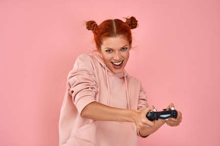 Smiling red-haired girl teenager playing with the joystick in a video game on a pink background....