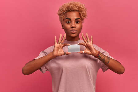 Happy young dark-skinned woman holding a bank card in front of her hands on a pink background. Girl...
