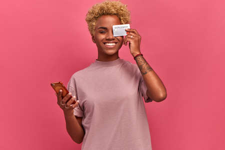 Happy young dark skinned freelancer woman holding smartphone and bank card, which closes one eye on pink background. Girl is happy that now she will receive good salary on card. Lifestyle and carefreeの写真素材