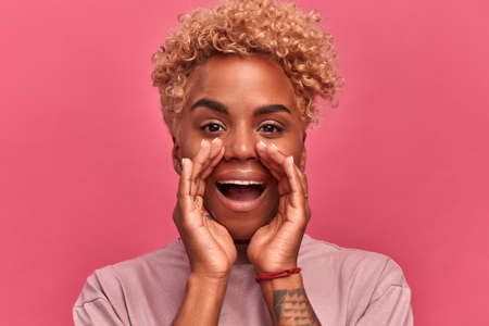 An African-American woman shouts into the camera and urges not to miss something important. Woman with short curly hair folded her arms around her mouth and shouted about crazy discounts. Close up.の写真素材