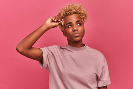 Brooding African American woman looks confused, thoughtful and distrustful against pink studio...