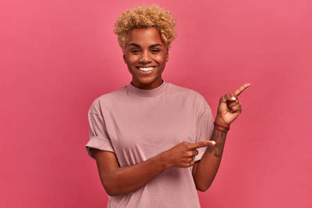 Attractive afro american woman smiles broadly at camera and points her fingers to the side, shows blank space for your promotion or advertisements on pink wall background. Positive human emotions.の写真素材