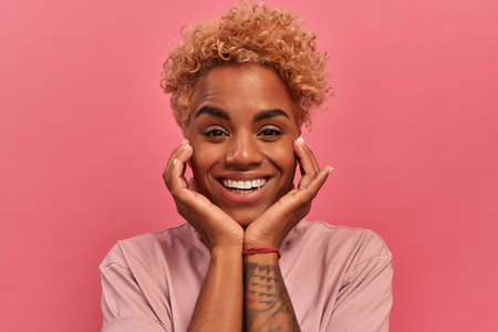 Portrait of pleasantly surprised dark skinned birthday female with blonde curly hairstyle touches...